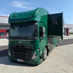 Grüner Volvo FM Pferdetransporter mit ausgefahrenem Wohnbereich, vor industrieller Kulisse.